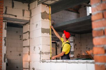 Construction worker at construction site measures the length of window opening and brick wall with tape measure. Cottage are made of porous concrete blocks, work clothes - jumpsuit and baseball cap - obrazy, fototapety, plakaty