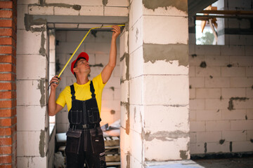 Fototapeta na wymiar Construction worker at construction site measures the length of window opening and brick wall with tape measure. Cottage are made of porous concrete blocks, work clothes - jumpsuit and baseball cap