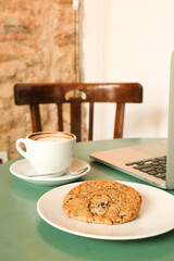 Remote working with laptop in a modern coffeeshop. Laptop, cup of coffee and chocolate chunk cookie lay on green table. No people.