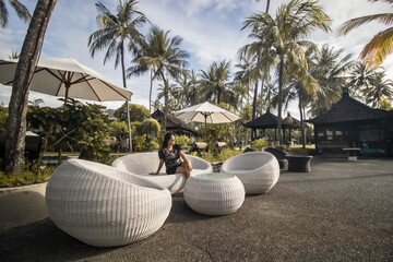 Pretty girl or woman with blond hair in hat drinks cocktail in sea beach cafe or bar under sun umbrella and palms on beautiful seascape