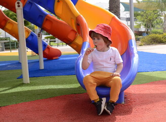 Fototapeta na wymiar View from above at little boy eating green lollipop candy outdoors on playground, looking up to camera. Sweet tooth. A boy in a red panama. Portrait of a child.