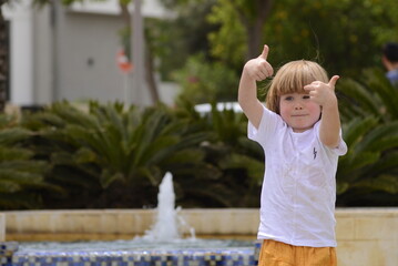 Naklejka premium A cute little boy is playing in the fountain. A child in yellow pants rejoices in summer and a sunny day. Walk with a child.