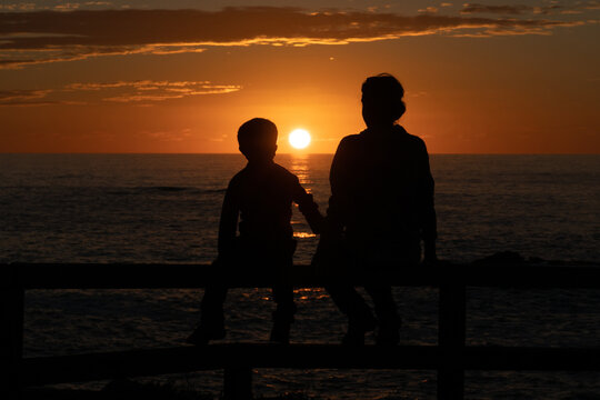 Mother and son at sunset looking at each other tenderly in front of the sea with the sun setting in the background and them silhouetted