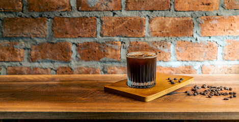 A Glass of ice coffee, cold americano in the morning over a red bricks wall background, and coffee beans on wooden counter bar table. Cold brew refreshment summer drink with copy space. Cafe Concept