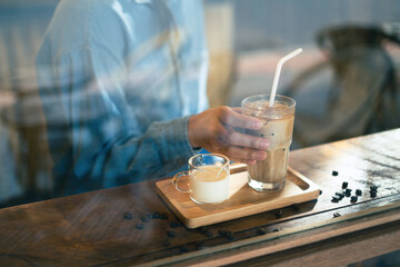 A hand holding a tall glass of iced latte coffee with milk on a wooden bar over a cafe glass window reflex at a Cafe coffee shop. Cold brew refreshment summer drink with copy space. Selective focus