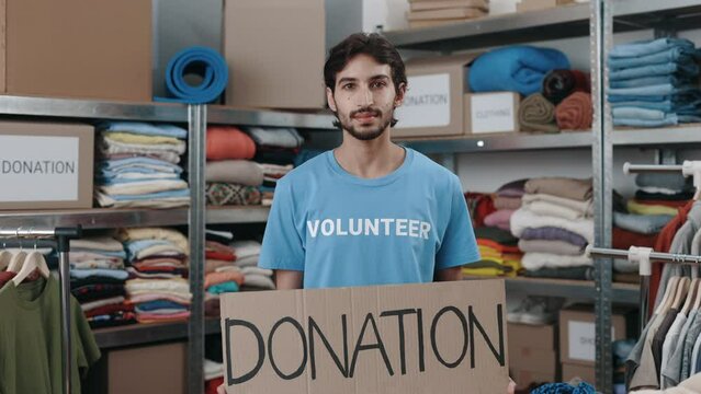 Portrait of energetic caucasian man looking at the camera and smiling while holding banner with donation word in charity shop. Volunteer working in organization and sorting donated clothes