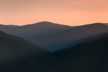Amazing landscape of mountains at sunrise. View of orange sky and hills covered forest at mist.
