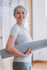 Happy fit middle aged old woman looking at camera, holding yoga mat, standing at home. Seniors...