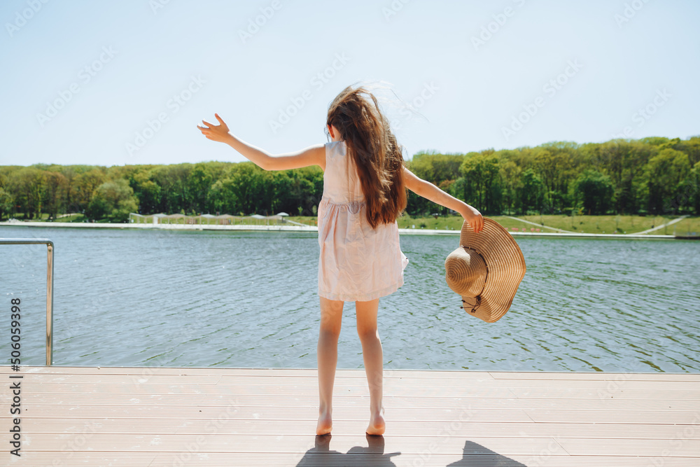 Wall mural a cute little girl is standing on the pier looking at the beautiful lake. child jumping and having f