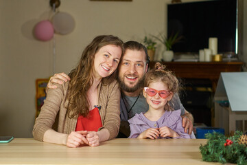 Happy family smiling portrait. A man and a woman hug their daughter. child has fun with his parents at the weekend at home at the table. Hugging is fun. Stylish girl in pink glasses expresses herself.