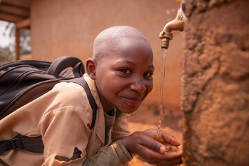 Black schoolchild collects water with his hands from the tap located in the schoolyard. Water...