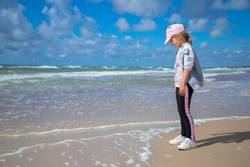 Happy child girl stands on the beach close to the sea and looks into the distance