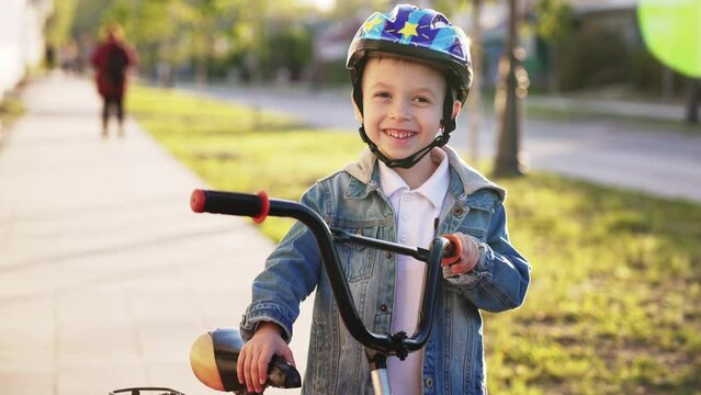 Happy smile child boy stands with bicycle in protective helmet for safe ride along on city street. Sports walk in nature on weekend. Healthy lifestyle. Process of learning to ride a bike in childhood