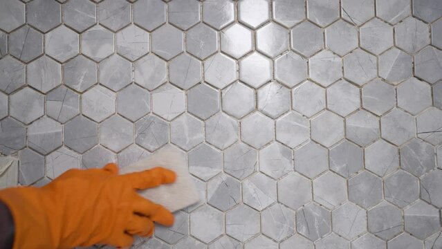 A man's hand in an orange rubber glove wipes the surface of a ceramic tile close-up. Stain removal, kitchen disinfection. House cleaning concept. Close-up of a gloved hand washes a wall tile.