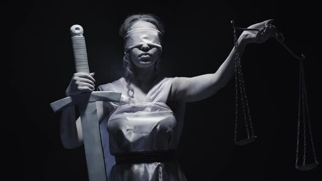Woman dressed and makeup as goddess justice holds sword and lifts scale, standing against dark background in studio, front view. Lady of justice raises scales as sign of triumph truth