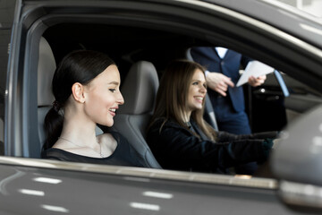 Gorgeous young girls sitting inside car, smiling and looking at camera. One of girls holding hands on steering wheel. Female customers of car dealership choosing auto in showroom.