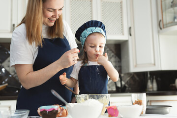 Cute little girl and her beautiful mom in matching aprons and caps play and laugh while kneading dough in the kitchen.