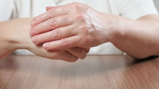 Elderly woman softly rubs moisturizing cream on palms with massage moves. Woman massage hard-working hands with nourishing cream sitting at table