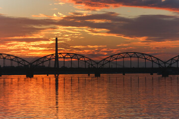 Beautiful sunset skies and their reflection in the calm waters of a wide river. Two bridge contours are visible.