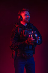 soldier in full gear with weapons. a man in headphones, a bulletproof vest, with a backpack and a belt drinks water from a can. red background. colored, blue-red light. world concept