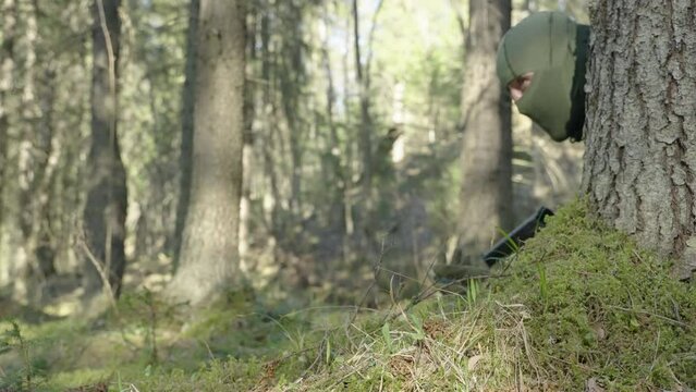 A soldier in military uniform and a mask aims at a target, looking out from behind a tree