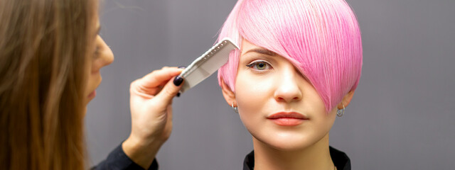 The female hairdresser is combing the dyed pink short hair of the young woman against the dark wall