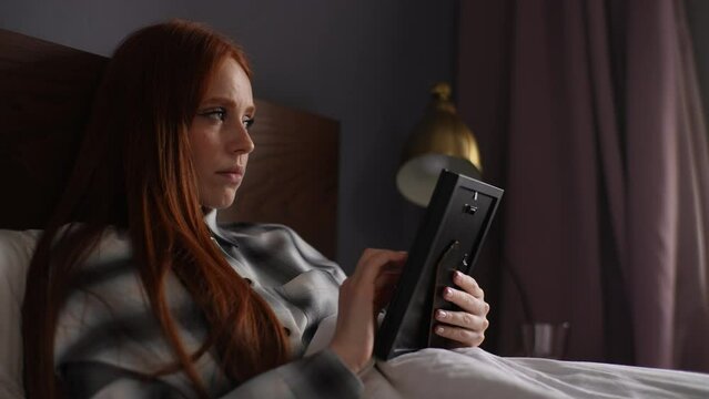Depressed young woman lying on bed in dark room holding photo in frame in hands and crying looking at it, touching picture with love. Sadness redhead female looking to photo with nostalgic feeling.