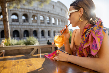 Woman drinking Spritz Aperol at outdoor cafe near Coliseum, the most famous landmark in Rome. Concept of italian lifestyle and traveling Italy. Caucasian woman wearing dress and shawl in hair - obrazy, fototapety, plakaty