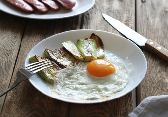 Fried eggs with fried zucchini in a plate on a wooden table. Behind is a plate of fried sausages.