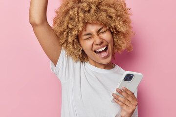 Excited cheerful young woman makes winner gesture keeps arm raised up holds mobile phone gets excellent news dressed in casual white t shirt isolated over pink background. Celebration concept