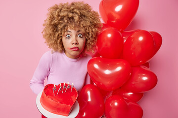 Impressed amazed woman celebrates special occasion holds delicious cake and bunch of red heart shaped balloons stares at camera has leaked makeup isolated over pink background. Holiday concept