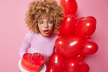 Shocked astonished European woman with curly hair stares impressed at camera cannot cannot believe in amazing news celebrates Valentines Day poses with tasty cake and bunch of heart balloons
