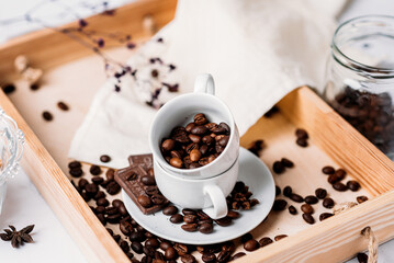 coffee beans in a white cup with a saucer, chocolate, anise, a cup of coffee on a wooden natural tray, morning drink in bed, light still life