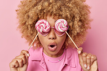 Breaking diet. Impressed curly haired young woman covers eyes with sweet spiral candies has mouth opened from amazement dressed in jacket isolated over pink background. Lollipops made for me