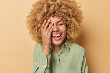 Happy cheerful woman makes face palm laughs joyfully expresses sincere feelings and positive emotions dressed in casual linen shirt isolated over brown background. Peope and happiness concept - obrazy, fototapety, plakaty