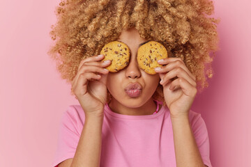 Portrait of culy haired young woman covers eyes with delicious cookies keeps lips folded dressed in...