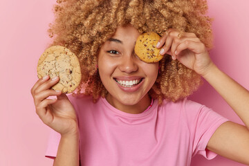 Positive carefree curly haired woman has sweet tooth holds delicious cookies over eye smiles pleasantly dressed in casual t shirt glad to eat delicious snack isolated over pink studio background.