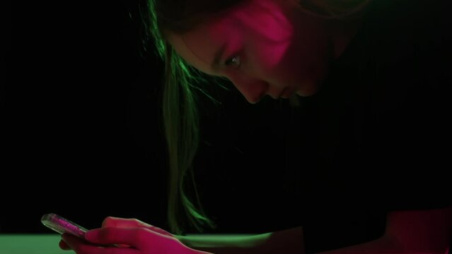 Girl read phone at dark night. Teenage girl using smart phone at night in purple and pink neon lights on black background. Reading message on phone. Side view. Close-up
