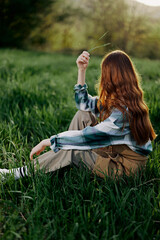 A woman sits on the green grass in a park with her back to the camera and relaxes in nature in the summer sunset evening light