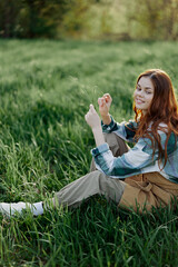 Fototapeta premium A woman sits on the green grass in a park with her back to the camera and relaxes in nature in the summer sunset evening light