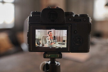 Horizontal close-up shot of camera recording video of stylish woman wearing headphones sitting at desk in loft room creating content for her blog