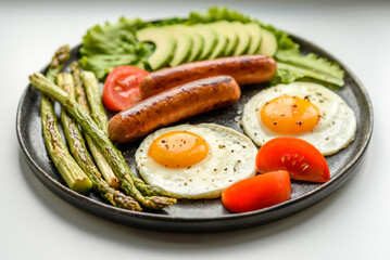 A delicious nourishing breakfast with fried eggs, sausages, asparagus, tomatoes, avocado, spices and herbs