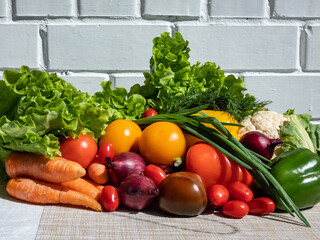 A pile of fresh, healthy and colorful vegetables in bright sunlight with white brick wall in background. Tomatoes, onions, carrots, peppers, cauliflower, salad, dill and greens