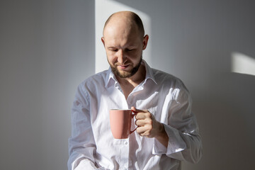 Smiling young man with a beard looks into a cup of coffee on a background of white wall.