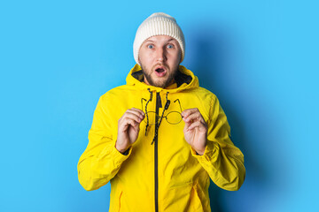Surprised young man in a yellow jacket holding glasses in his hands on a blue background