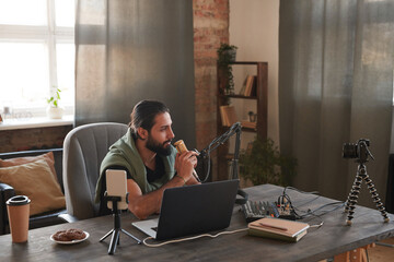 Horizontal shot of handsome Middle Eastern man sitting at table in front of laptop setting microphone to start online stream