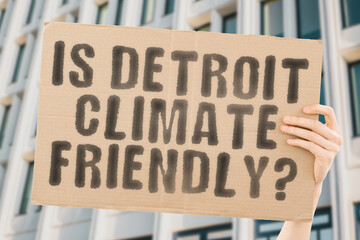 The question " Is Detroit climate-friendly? " is on a banner in men's hands with blurred background. Support. Team. Activist. Urban. Sunset. Carbon. Ecology. Energy. New. Clean. Warming. Waste