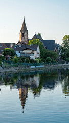 Fototapeta na wymiar The church tower and other village buildings reflect in the placid waters of the Rhine River in the early evening.