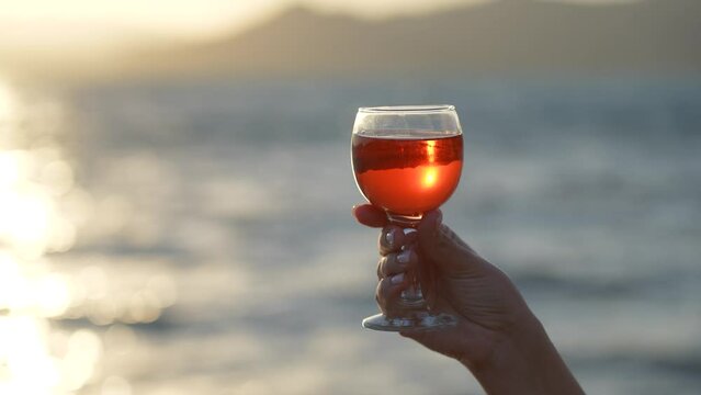 Closeup view 4k stock video footage of female hand holding glass of rose wine isolated on blurry sunset sea beach background. Woman relaxing on sunny beach outside. Happy beach party or dating concept