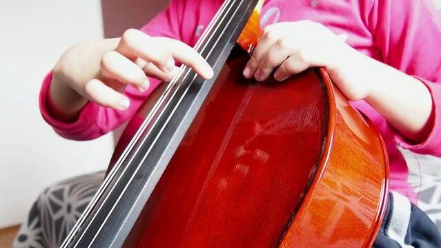 Picicato on the cello. A child girl in a pink jacket plays pizzicato with her fingers the cello without a bow. An entry-level violoncello study at a music school.